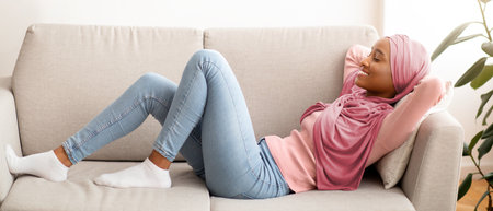 Beautiful black woman in hijab lying on sofa with closed eyes, taking break and relaxing at home, copy spaceの写真素材