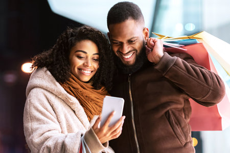 Portrait of happy afro couple using smartphone with shopping bagsの写真素材