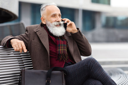 Senior man talking on phone, sitting on bench by officeの写真素材