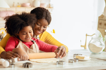Happy black mom and her daughter rolling up dough together in kitchenの写真素材