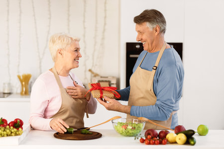 Senior Man Giving Birthday Gift To Wife Standing In Kitchenの写真素材