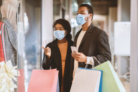 Happy young african american couple of shoppers in protective mask walk in mall, hold colorful bags