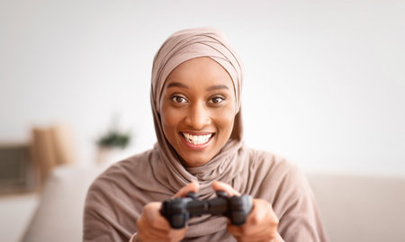 Portrait of happy Muslim woman in hijab holding joystick, playing videogame at homeの写真素材