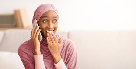 Modern communications concept. Surprised Muslim woman in hijab talking on cellphone at home, chatting online with friendの写真素材
