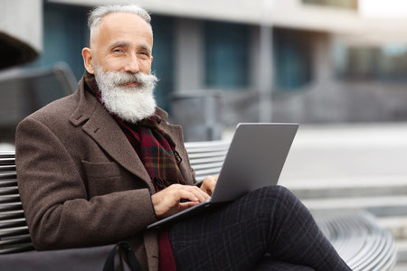 Wealthy grey-haired aged man sitting by office building, using laptopの写真素材