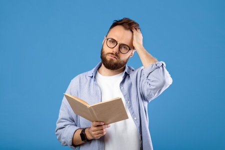 Studying is hard. Young bearded guy having problem understanding textbook on blue studio background