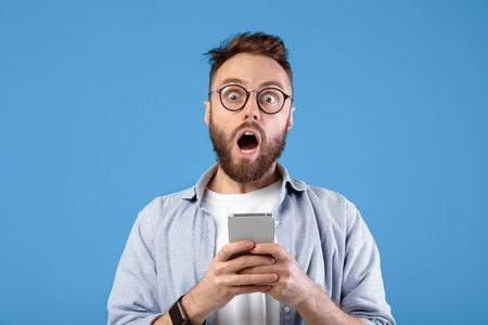 Shocked bearded guy staring at mobile phone in disbelief over blue studio background
