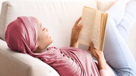 Cozy stay at home activities. Black young woman in hijab reading her favorite book, lying on sofa in...