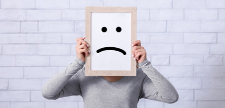 Depression, sadness, negative emotions concept. Young woman holding picture frame with sad emoticon...