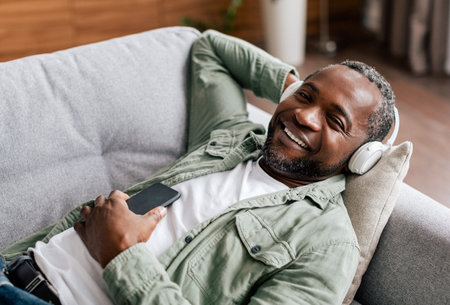 Satisfied middle aged african american man in casual, in wireless headphones enjoys favorite music with phoneの写真素材