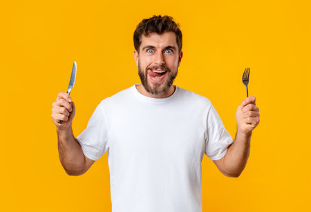 Hungry young guy holding knife and fork on yellow background