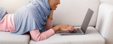 Side view of black woman in hijab using laptop with blank screen on sofa at home, copy space