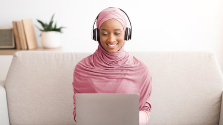 Muslim businesswoman in hijab and headset having video call on laptop at home office, talking with...