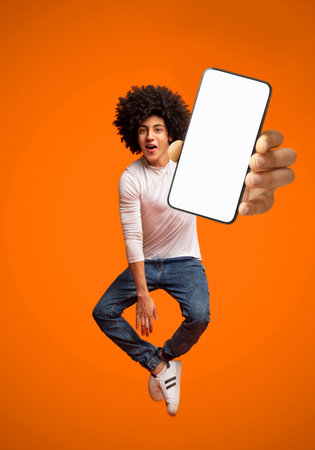 Funny young black teen guy jumping on air, showing cellphone with white empty screen on orange studio background, mockupの写真素材