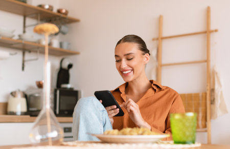 Excited lady using smartphone in kitchen while having dinner, sitting at table and texting on phone, free spaceの写真素材