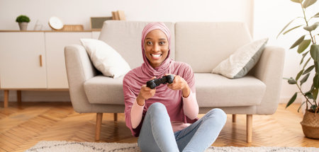 Happy Muslim woman in hijab sitting on floor with joystick, playing videogame at home, full length...