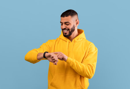 Young arab guy checking his smartwatch on blue studio background. Happy middle eastern man using fitness trackerの写真素材