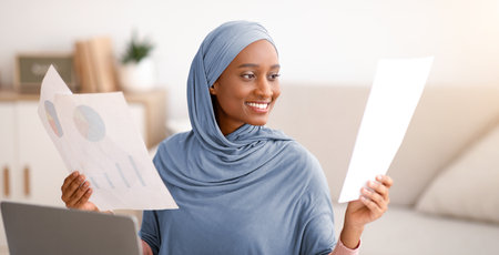 Work from home concept. Happy black Muslim woman in hijab looking through documents in front of laptop indoorsの写真素材