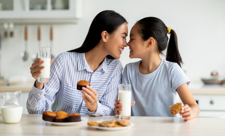 Loving asian mother and little daughter having snacks and drinking milk in kitchen, enjoying freshly baked cakesの写真素材