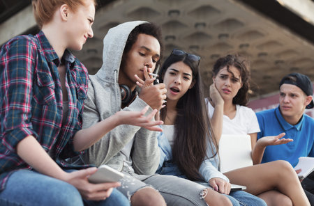 Black teen guy smoking cigarette, his friends judging himの写真素材