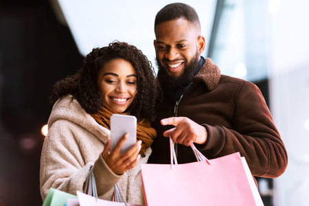 Portrait of happy black couple using phone with shopping bagsの写真素材