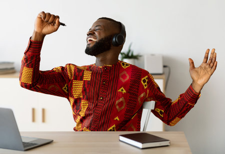 Break In Work. Cheerful African Guy In Traditional Shirt Singing At Workplaceの写真素材
