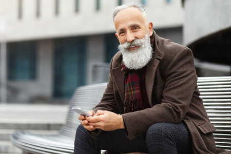 Smiling senior grey-haired man with mobile phone sitting on benchの写真素材