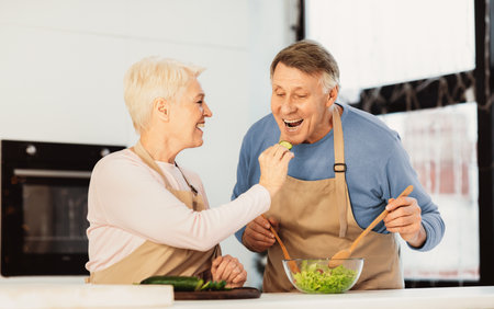Senior Family Couple Feeding Each Other Cooking Dinner In Kitchenの写真素材