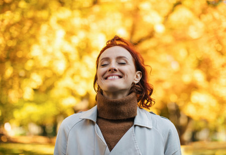 Satisfied caucasian millennial red-haired female in raincoat enjoys warm autumn and freedomの写真素材