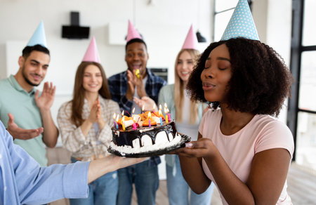 Black lady blowing candles on birthday cake, closing eyes, making wish, celebrating birthday with her diverse friendsの写真素材