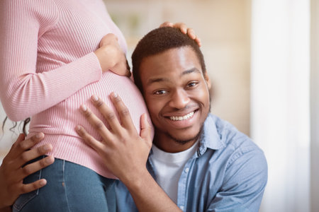 Handsome black man listening his wifes pregnant belly at homeの写真素材