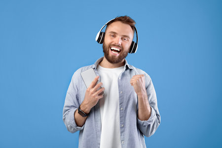 Handsome man with cellphone listening to music in headphones, singing along to his favorite song on blue backgroundの写真素材