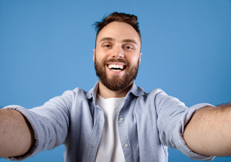 Joyful bearded guy taking selfie on blue studio backgroundの写真素材