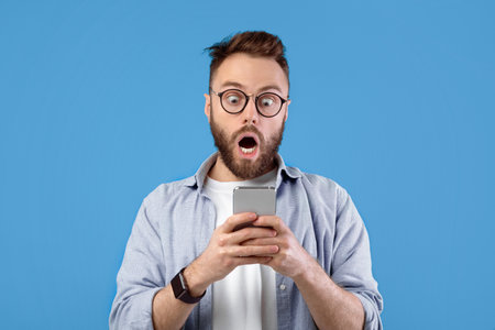 Shocked Caucasian guy staring at cellphone with open mouth over blue studio background, panoramaの写真素材