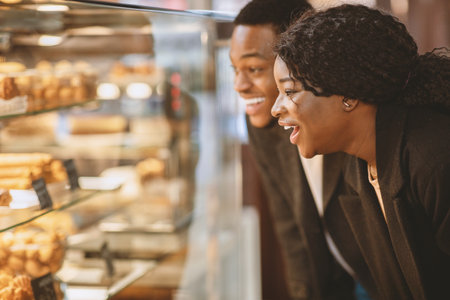 Surprised happy millennial woman and man standing in front of glass showcase with pastries and cakesの写真素材