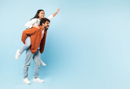 indian man giving piggyback ride to wife against blue backdropの写真素材