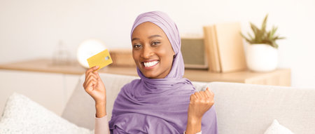 Happy black woman in hijab holding credit card, making YES gesture, shopping online on laptop computer at homeの写真素材