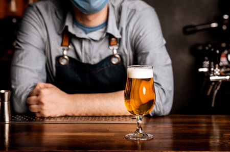 Bartender in protective mask leans on bar counter on which stands glass of light beer with foamの写真素材