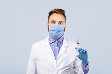 Immunization. Young male doctor wearing protection, holding syringe with coronavirus vaccine over grey backgroundの写真素材