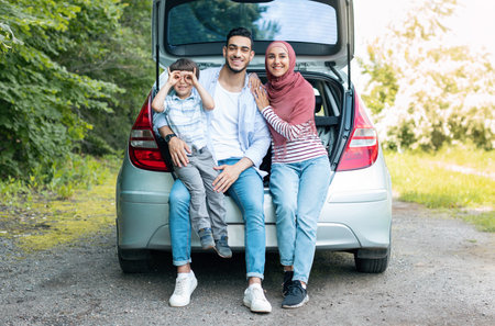 Smiling young middle eastern man and wife in hijab holding baby, sitting in car trunk, resting from trip outdoorの写真素材