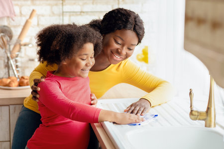 Happy Black Mom Teaching Her Cute Little Daughter Housekeeping, Cleaning Kitchen Togetherの写真素材
