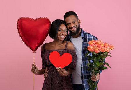 Valentines Day. Young black couple with red heart, balloon and bouquet of roses posing on pink studio backgroundの写真素材