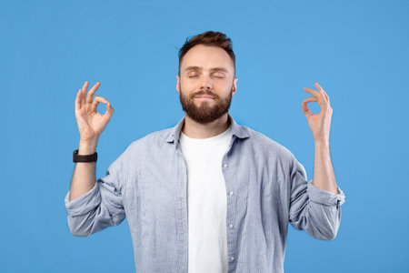 Stress management. Peaceful young guy meditating with closed eyes, making gyan mudra with his hands on blue backgroundの写真素材