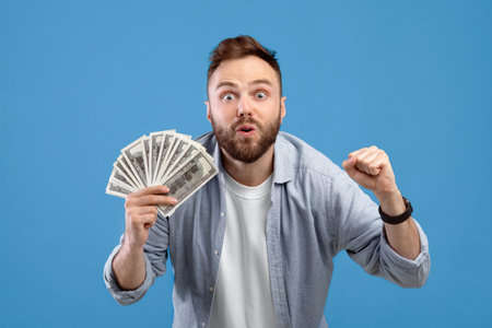 Excited millennial man holding money, gesturing YES over big win on blue studio background