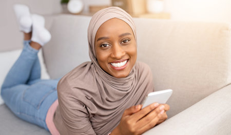 Online life. Smiling black woman in hijab lying on sofa with smartphone, chatting or browsing web at...