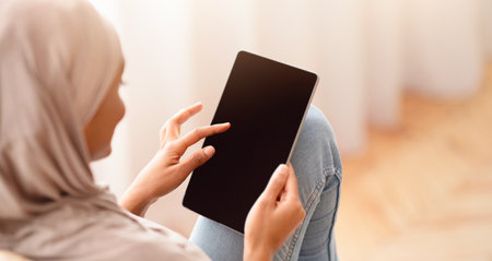 Black Arab woman in hijab using tablet with empty screen while sitting in cozy armchair at home,...