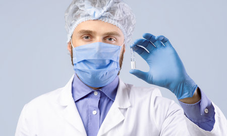 Vaccination and health protection. Male doctor in facial mask holding vaccine or medication on grey...