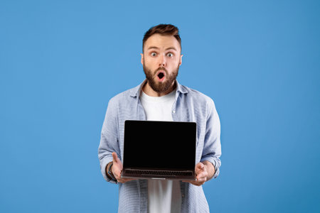 Shocked bearded guy showing laptop with blank screen on blue studio background, mockup for your...