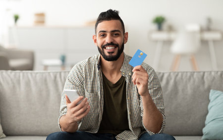 Ordering goods online. Happy young Arab guy with credit card and smartphone shopping on web at homeの写真素材