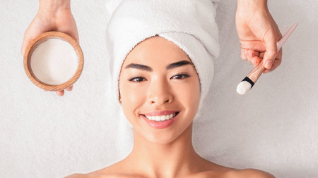 Facial Treatment. Cosmetologist applying clay mask on asian womans face at spaの写真素材